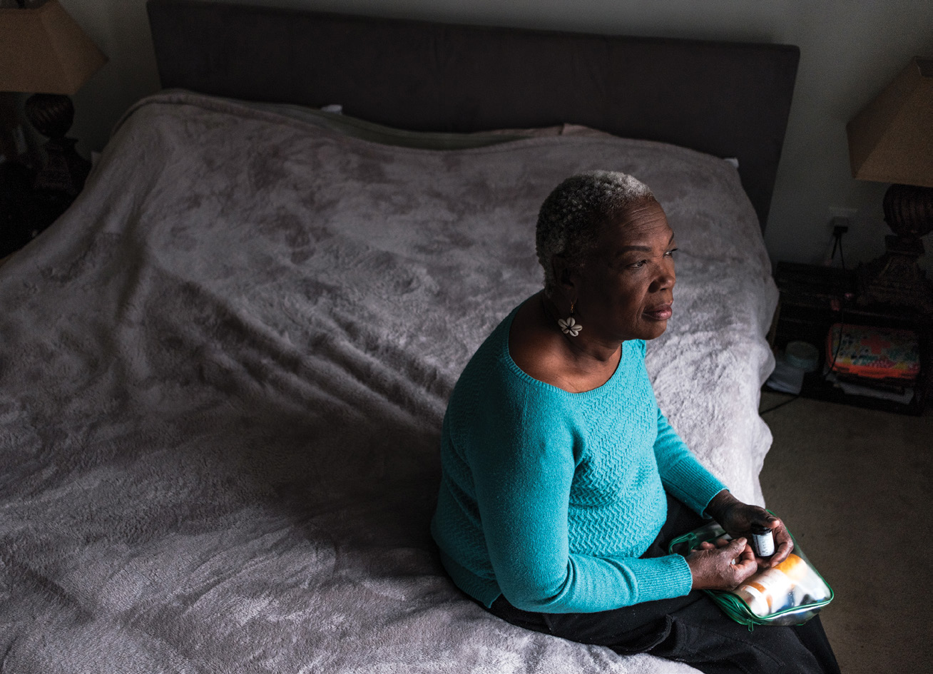 an older woman with graying hair sits on her bed, holding a bottle of prescription pills and a bag of other medicines
