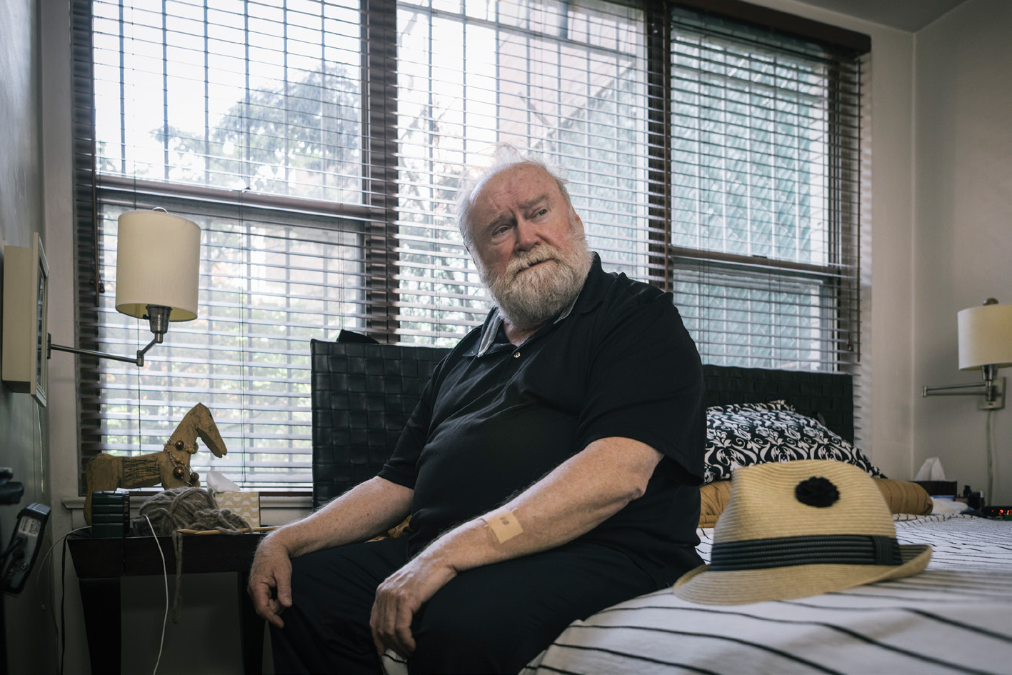 an older man with gray hair and a small bandage on his arm sits on a bed by the window