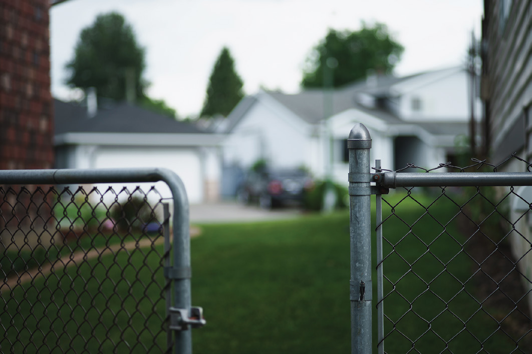 a chain link fence gate opens to a grassy yard and a white one-story suburban house with a few trees in the distance
