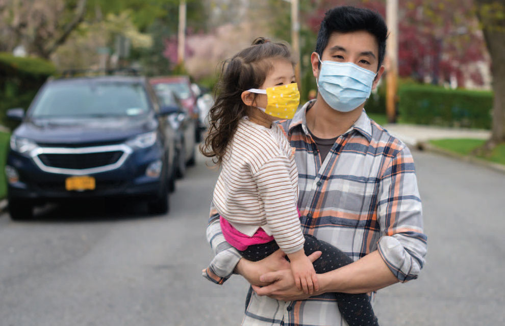 Standing in a tree-lined neighborhood street, a father wearing a blue surgical face mask holds his young daughter who is wearing a yellow cloth mask.
