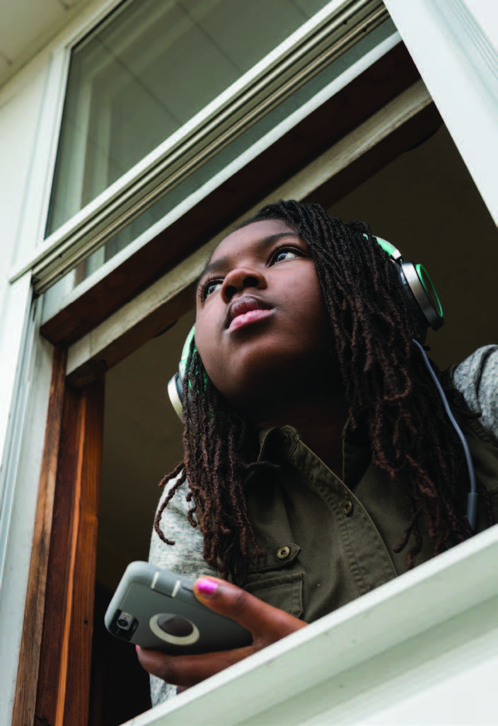 A teenage girl, wearing headphones and holding a mobile device, rests against her open window and looks out to the distance.