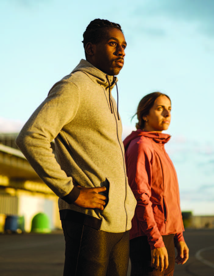 Two young adults in athletic clothing stand slightly separated from one another next to an outdoor running track.