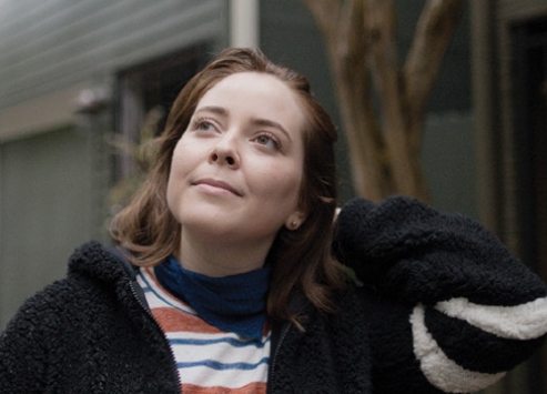 A young woman wearing a black sweater and striped t-shirt stands outside by an apartment building looking up toward the sky.